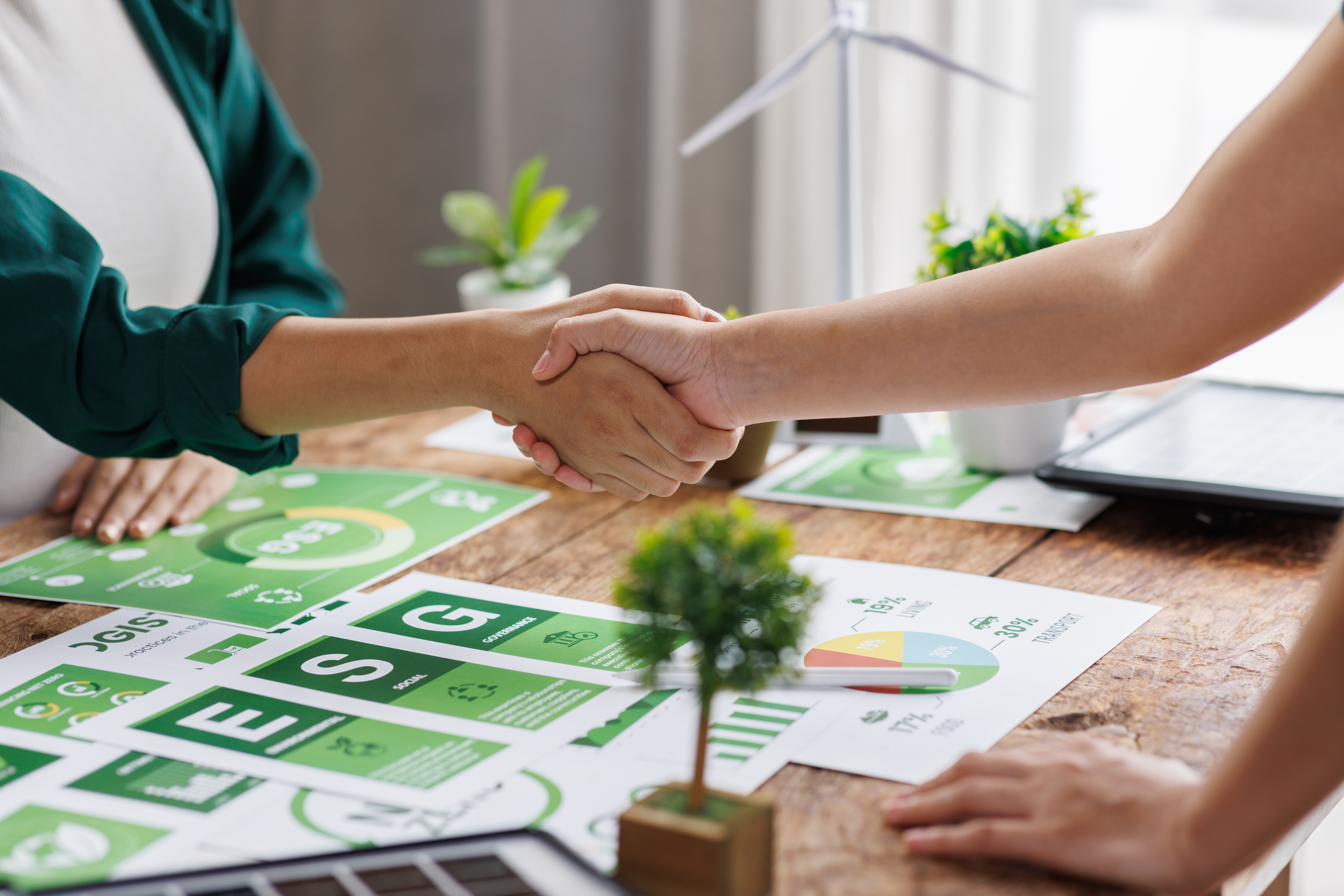 Two people shake hands over a desk with infographics and small potted plants on it.