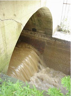 Muddy water exiting a round opening in a concrete headwall into a concrete channel.