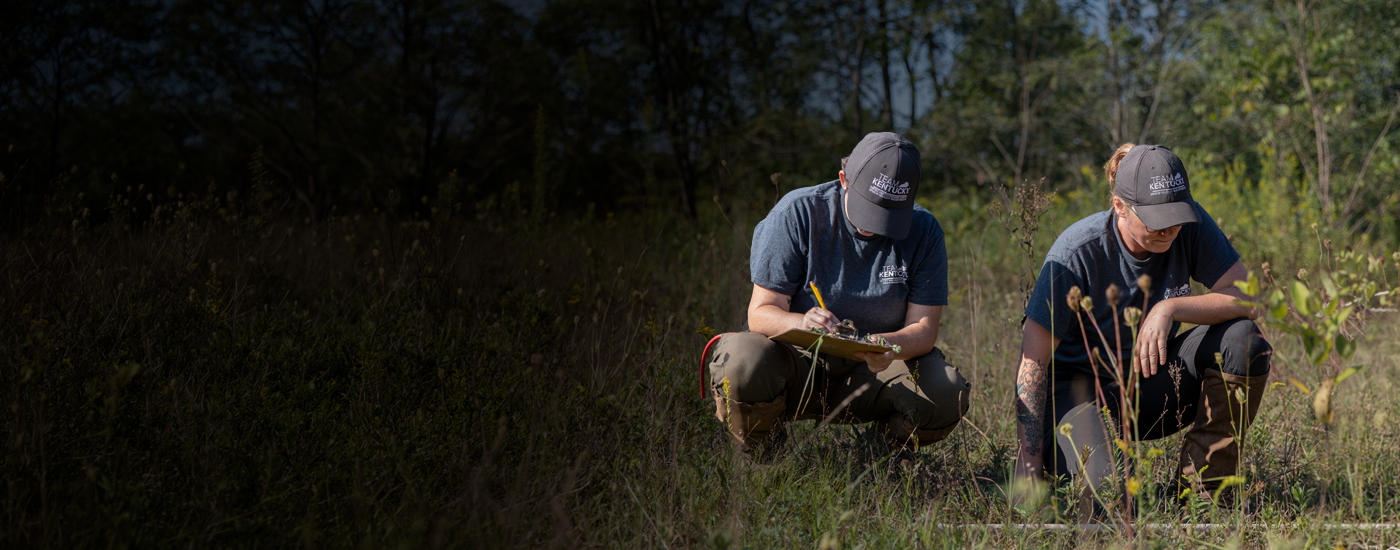 Two people wearing Team Ky hats in a field. One is taking notes